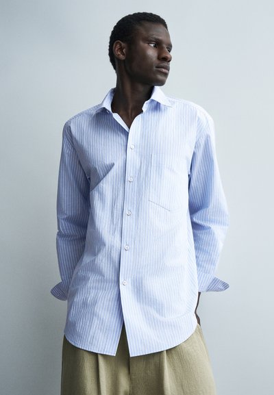 Jeune homme regardant vers la droite, portant une chemise à rayures bleu clair et blanc boutonnée et un pantalon beige, debout devant un fond gris clair uni.