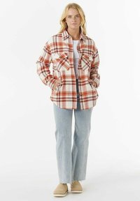 Frau in einer rot-weiß karierte Jacke über einem weißen Shirt, hellblauen Jeans und beigen Slip-On-Schuhen, die mit Händen in den Jackentaschen steht.