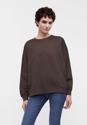 Unge med kort krøllet hår iført en løs mørkebrun sweatshirt og blå jeans, stående mod en ensfarvet lys baggrund.