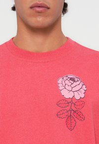 Camiseta de algodón color coral con un gran gráfico de rosa rosa en el lado izquierdo del pecho, contorneada en negro, con hojas detalladas debajo.