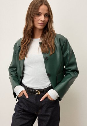 Femeie cu păr lung și castaniu, purtând o jachetă verde din piele, bluză albă cu mâneci lungi, curea neagră și blugi din denim închis, cu mâinile în buzunare.