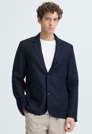 Junger Mann mit lockigem Haar, der einen dunkelblauen Blazer über einem weißen Hemd und beigen Hosen trägt, vor hellem Hintergrund stehend.