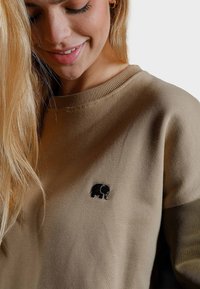 Felpa khaki con colletto rotondo e polsini a coste, decorata con un piccolo elefante nero ricamato sul petto. Tessuto morbido.