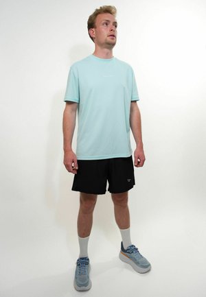 Jeune homme debout, portant un t-shirt bleu clair, un short noir, des chaussettes blanches et des chaussures de course grises sur un fond blanc uni.