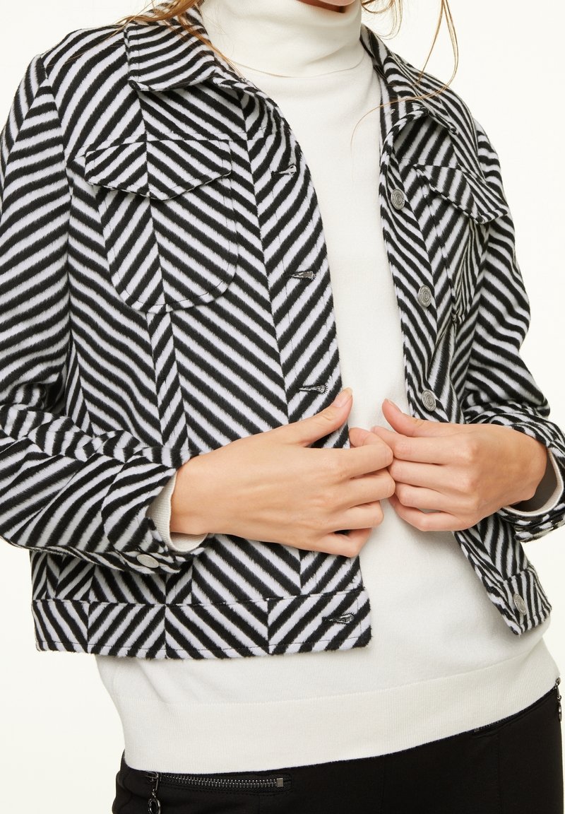 Sort og hvid stribet jakke med to frontlommer, knapdetaljer og cropped design, lagdelt over en hvid turtleneck sweater.