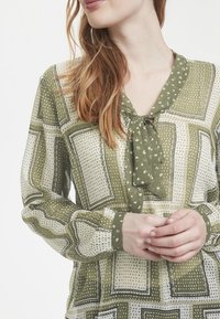 Zelená blůza se vzorem, s výstřihem do V a zavazováním. Obsahuje geometrické vzory a puntíky, vyrobena z lehkého materiálu s dlouhými rukávy.