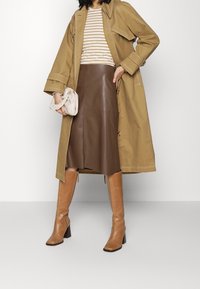 Beige Trenchcoat mit großem Kragen, gestreiftem Oberteil, brauem Lederrock und knielangen sandfarbenen Stiefeln. Kleine cremefarbene Handtasche in der Hand.