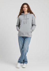 Felpa grigia con testo nero "SPOSIAMOCI", tasca a marsupio, cappuccio regolabile con cordino, abbinata a jeans blu e scarpe da ginnastica bianche.