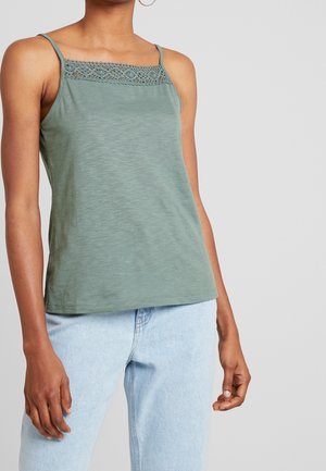 Kvinde iført ærmeløs grøn camisole med blondekant ved halsudskæringen og lyseblå denimjeans mod en ensfarvet baggrund.