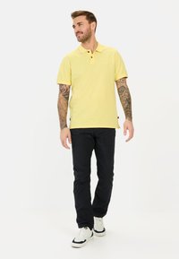 Gelbes Polohemd mit Kragen, kurzen Ärmeln und zwei Knöpfen; kombiniert mit dunklen Hosen und weißen Sneakers. Tätowierte Arme sichtbar.