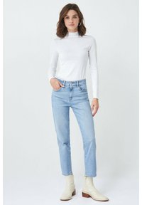 Suéter blanco de manga larga con cuello alto, jeans rectos de mezclilla azul claro y botines crema con tacón apilado, de pie contra un fondo sencillo.