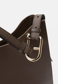 Καφέ δερμάτινη τσάντα χειρός με υφασμάτινη υφή, χρυσές λεπτομέρειες και ιμάντα με ρυθμιζόμενες τρύπες για ευέλικτες επιλογές μεταφοράς.