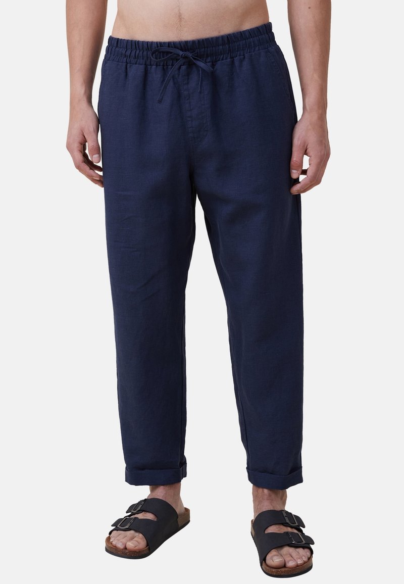 Homme portant un pantalon bleu marine à cordon de serrage avec les ourlets repliés aux chevilles, et des sandales noires à double bride, sur un fond uni.