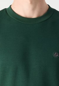 Felpa verde a maniche corte con colletto a girocollo a coste e piccolo marchio nero sul lato sinistro del petto, realizzata in morbido tessuto di cotone.