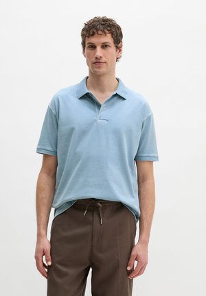 Junger Mann mit kurzen lockigen braunen Haaren, der ein hellblaues Poloshirt und braune Hosen mit Kordelzug trägt, vor einem schlichten weißen Hintergrund.