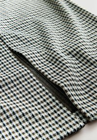 Svart, vitt och brunt houndstooth-mönstrat tyg med en slät yta, försett med en dekorativ dragkedja på ena sidan.