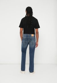 Jeans de ganga azul-escuro com pernas retas e bolsos traseiros. Usados com uma t-shirt preta e sapatos de cor clara. Apresenta um emblema de marca na cintura.