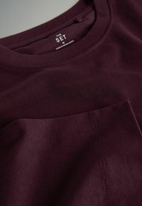 Bordowa koszulka z miękkiej bawełny, z okrągłym dekoltem, krótkimi rękawami oraz kieszenią naszywaną na lewej piersi, z przeszyciami.