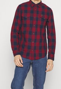 Rød og marineblå plaid flannel skjorte med knap-ned krave, to brystlommer og frontknapper. Stoffet har et blødt, tekstureret udseende.