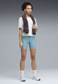 Ελαφρύ λευκό τοπ με κοντά μανίκια, στενά σορτς σε γαλάζια απόχρωση, γκρι πετσέτα περασμένη στο λαιμό, λευκά αθλητικά παπούτσια με τυρκουάζ λεπτομέρειες, κότσος με σγουρά μαλλιά.
