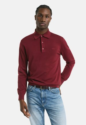 Mann trägt ein kastanienbraunes Langarm-Poloshirt, das in eine hellblaue Jeans mit einem braunen Gürtel gesteckt ist, und steht vor einem einfarbigen Hintergrund.