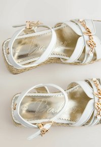 Witte leren sleehakken met gouden kettingdetail, enkelbandjes en gevlochten jutezolen, van bovenaf gezien op een beige achtergrond.