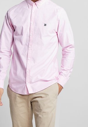 Muž v světle růžové košili na knoflíky s malým černým logem "H" na hrudi a v béžových kalhotách, ruka v kapse, na šedém pozadí.