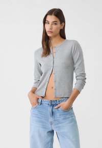 Kvinna med långt brunt hår bär en oförknäppt grå cardigan och ljusblå jeans, står med händerna i fickorna mot en vit bakgrund.