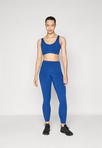 Top desportivo azul e leggings de cintura alta feitos de material elástico, com design sem costuras e bolsos laterais, combinados com ténis pretos.