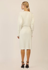 Weißes Rippstrickkleid mit enger Passform und langen Ärmeln. Der Saum endet auf halber Wade, kombiniert mit schwarzen Sandalen mit Absatz und Riemchen.