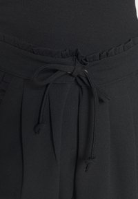 Pantalones de tela negra con cintura fruncida, pliegues frontales y un lazo de cordón para abrochar. Textura suave con un detalle de diseño sutil.