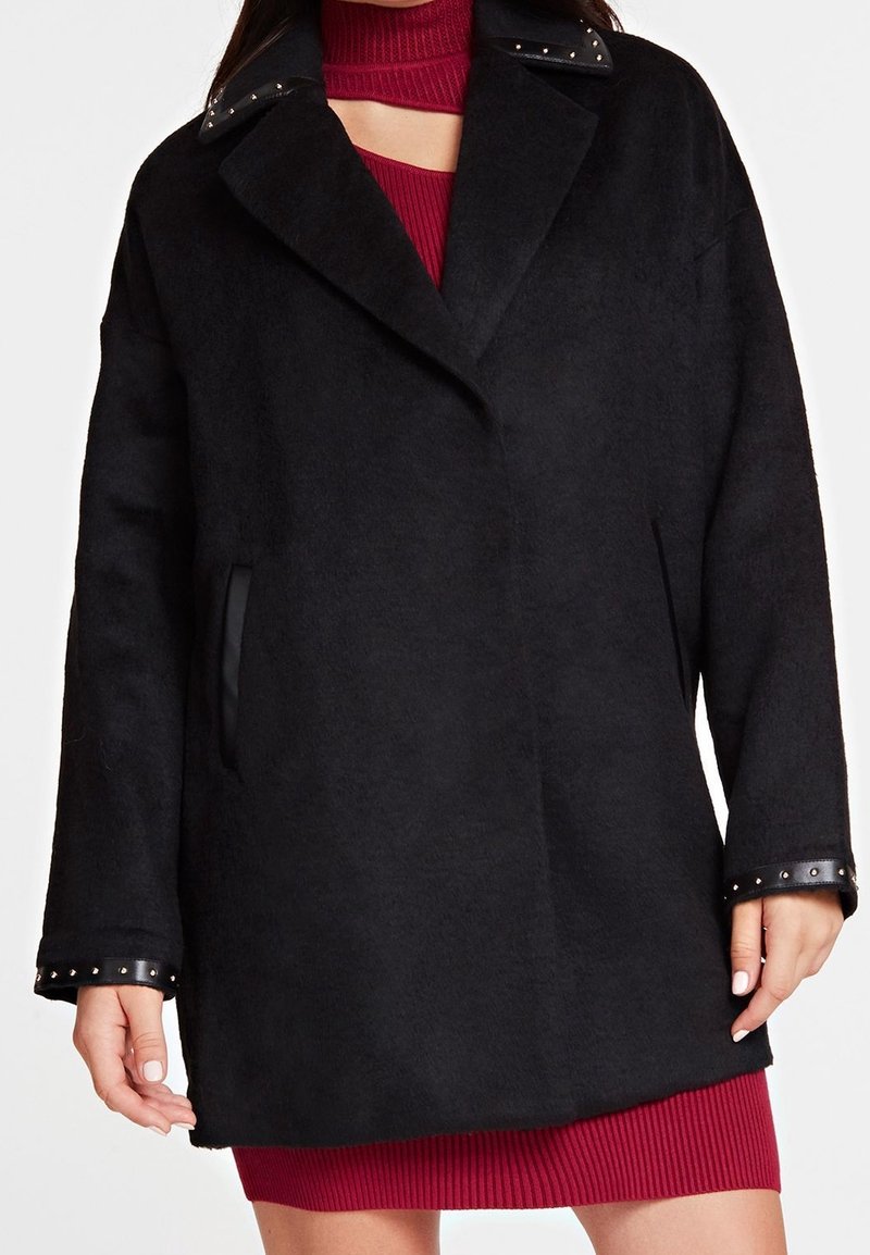 Femme portant un manteau en laine noir avec des garnitures en cuir clouté au niveau du col et des poignets, par-dessus une robe rouge en maille côtelée.