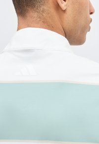 Gros plan sur le cou et l'épaule d'une personne portant une chemise blanche et bleu clair avec un logo Adidas subtil au niveau du col arrière.