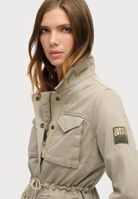Veste kaki avec un col haut, fermeture éclair, deux poches avant et taille ajustable par cordon. Caractérisée par un patch de marque vert sur la manche.