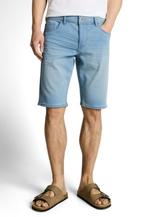 Man draagt lichtblauwe denimshorts, wit overhemd en bruine sandalen met gesp, staat met één hand in de zak.
