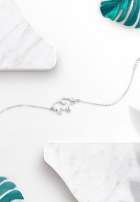 Braccialetto a forma di elefante in argento su una delicata catena, con finitura lucida; esposto su una superficie in marmo bianco e foglie verdi.