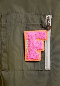 Tkanina w oliwkowym kolorze z naszywaną kieszenią i beżowym zamkiem błyskawicznym, z różową, puszystą łatką litery "F" na pomarańczowym filcu przymocowaną do kieszeni.