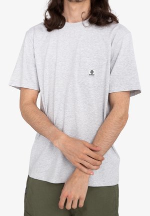 Homme portant un t-shirt à manches courtes gris clair avec une poche avant et le logo de la marque Element, associé à un pantalon vert olive, debout les mains jointes.