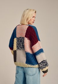 Knit sweater featuring a patchwork design with multiple colors, including red, blue, yellow, and pink. Textured, oversized fit.