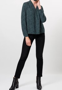 Blouse à motifs verts avec un décolleté en V profond, des manches longues et des épaules froncées, assortie à un jean slim noir et des bottines.