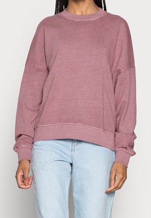 Femme portant un sweat-shirt ample à manches longues rose poussiéreux et un jean bleu clair, debout devant un fond uni.