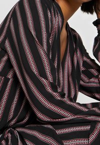 Černá tkanina s vertikálními pruhy v červené a bílé barvě. Disponuje uvolněným designem s hlubokým výstřihem a volnými rukávy.