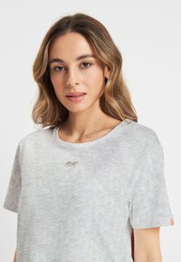 Vrouw met lichtbruin haar die een lichtgrijze T-shirt draagt met een klein hartontwerp nabij de halslijn, tegen een effen witte achtergrond.