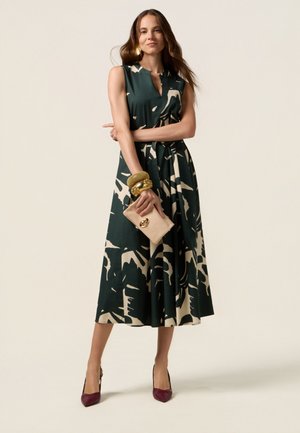 Ärmelloses Kleid in Dunkelgrün mit cremefarbenem Blumenmuster, tailliert, aus fließendem Stoff und knielang. Die Accessoires umfassen goldene Armbänder und eine Clutch.
