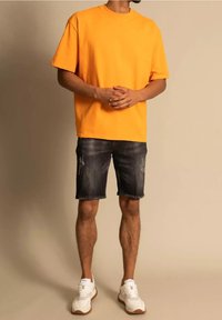 Oranssi puuvillainen lyhythihainen t-paita yhdistettynä kuluneisiin tummiin farkkushortseihin. Valkoiset urheilukengät täydentävät asun.