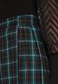 Gonna a quadri in verde scuro e nero con linee gialle e bianche; presenta pieghe e una parte superiore nera testurizzata con un motivo a zigzag.