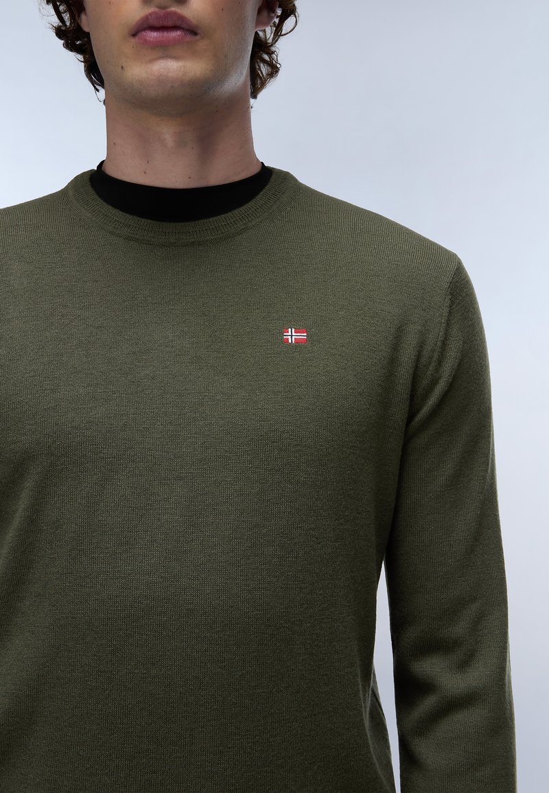 Maglione verde oliva con scollatura rotonda, lavorazione a maglia testurizzata e un piccolo emblema della bandiera norvegese rosso e bianco sul petto.