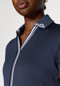 Námořnická polokošile s pruhovaným límcem a lemy, vyrobená z hladkého materiálu s mírným leskem; zvýrazněná předním zipem.