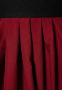 Gonna plissettata rossa con un tessuto strutturato, dotata di un elastico nero in vita. Le pieghe sono profonde e uniformemente distanziate, creando un aspetto ben definito.