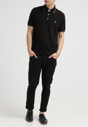 Polo noir à manches courtes, col et poignets contrastés avec des détails blancs, logo sur la poitrine ; associé à un pantalon et des chaussures noirs.